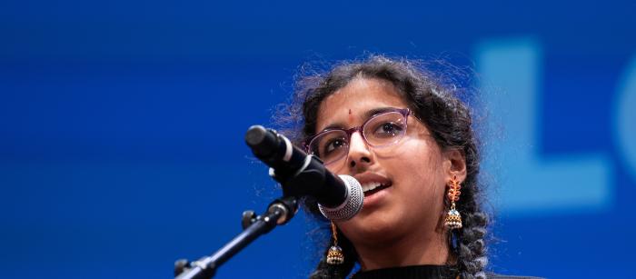 A young woman with glasses and two long braids speaks into a microphone