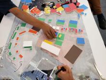 An overhead camera shot of hands from two individuals working on a project with colored tiles and a map 