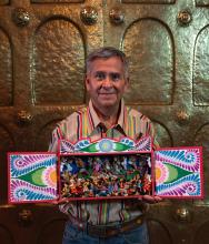 A man with grey hair stands holding a display of colorful figurines set in a hand-painted Nativity box