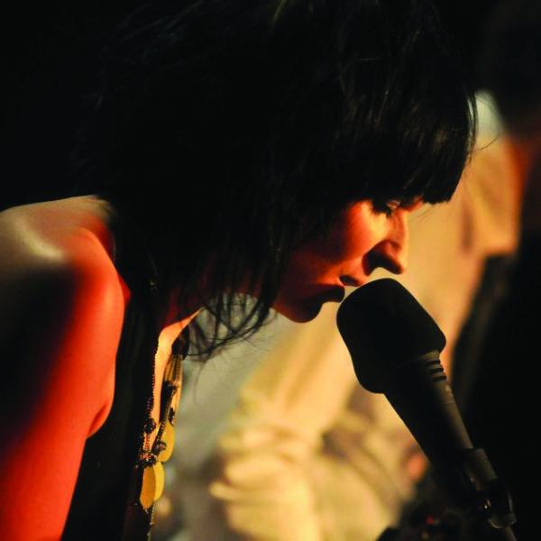 Woman with short black hair singing into a mic with a man out of focus in the background.